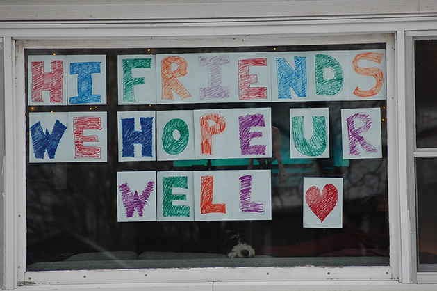 A dog sits in a window below a sign reading "Hi Friends We Hope U R Well"
